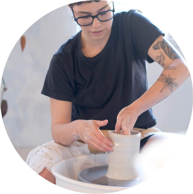 A person with short dark hair, glasses, and tattoos on their arm is shaping a clay pot on a pottery wheel in a bright ceramics studio. They are wearing a black t-shirt and white pants, focusing intently as they mold the clay. The setting is clean and minimal, emphasizing the hands-on experience of wheel-throwing in a Vancouver pottery class.