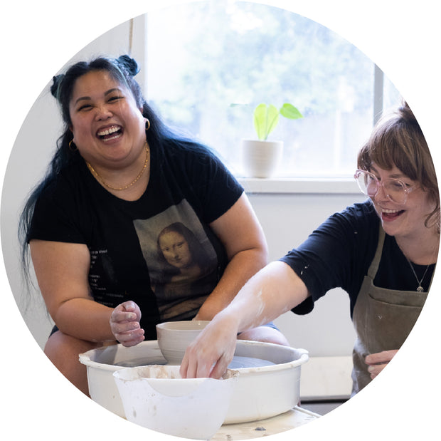 Two smiling participants enjoying a hands-on pottery workshop in Vancouver, shaping clay on a pottery wheel in a bright and welcoming ceramics studio.